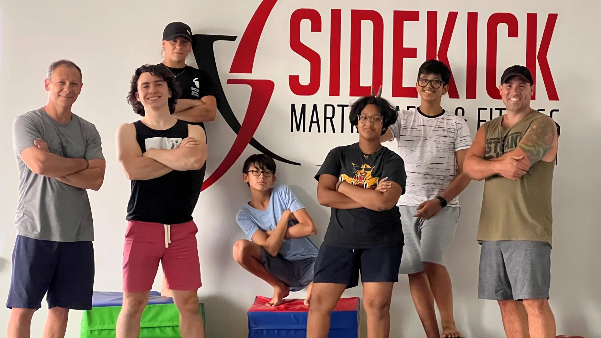 Teen and adult Sidekick students standing together in front of the Sidekick Martial Arts & Fitness logo